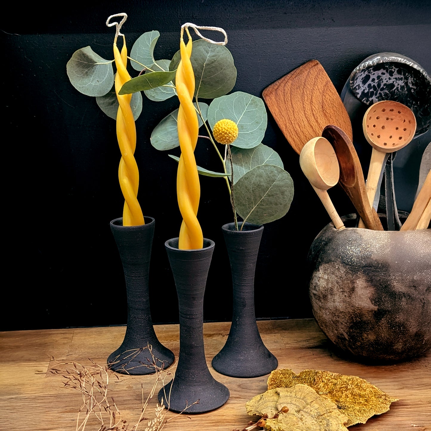 Three black clay taper holders with Twin Flame beeswax candles and eucalyptus stems arranged beside wooden kitchen utensils—evoking a modern, organic home setting.