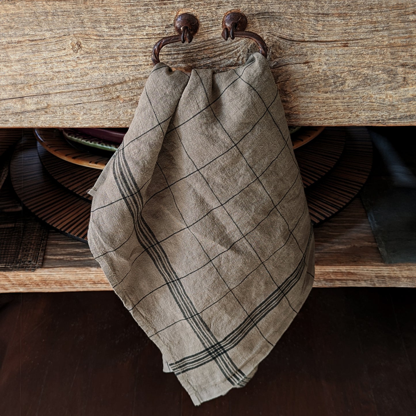 A simple classic plaid pattern linen kitchen towel hangs neatly from cast iron hooks on a weathered wooden shelf.