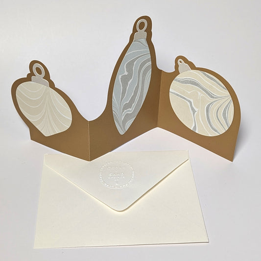 A gold trifold ornament notecard standing open on a white surface, featuring three Suminagashi-marbled ornament panels in soft neutral tones with an embossed CEDAR Y SAND envelope below.