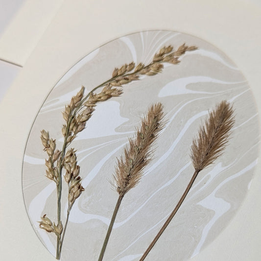 Close-up of Walking Dunes Straw pressed flora notecard with two feathery golden grass plumes and an arching seed stem over hand-marbled Suminagashi paper.