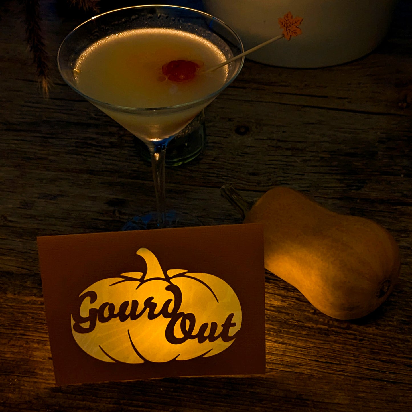 Gourd Out Notecard illuminated from within by an Autumn Glow Tealight, glowing softly beside a cocktail and butternut squash on a rustic wood table.