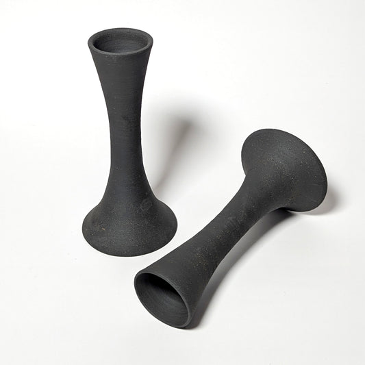 Two handcrafted black clay candle taper holders displayed on a white background. One stands upright while the other lies horizontally, revealing smooth throwing rings and organic curves.