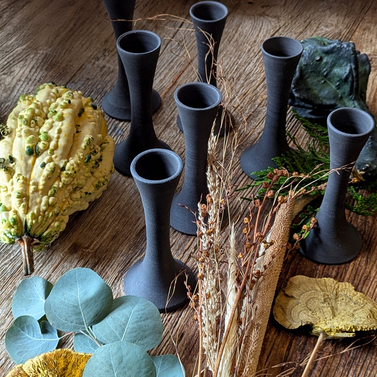 Group of black clay candle taper holders arranged on a rustic wooden surface surrounded by gourds, cedar, and dried botanicals, evoking a warm autumnal tablescape.
