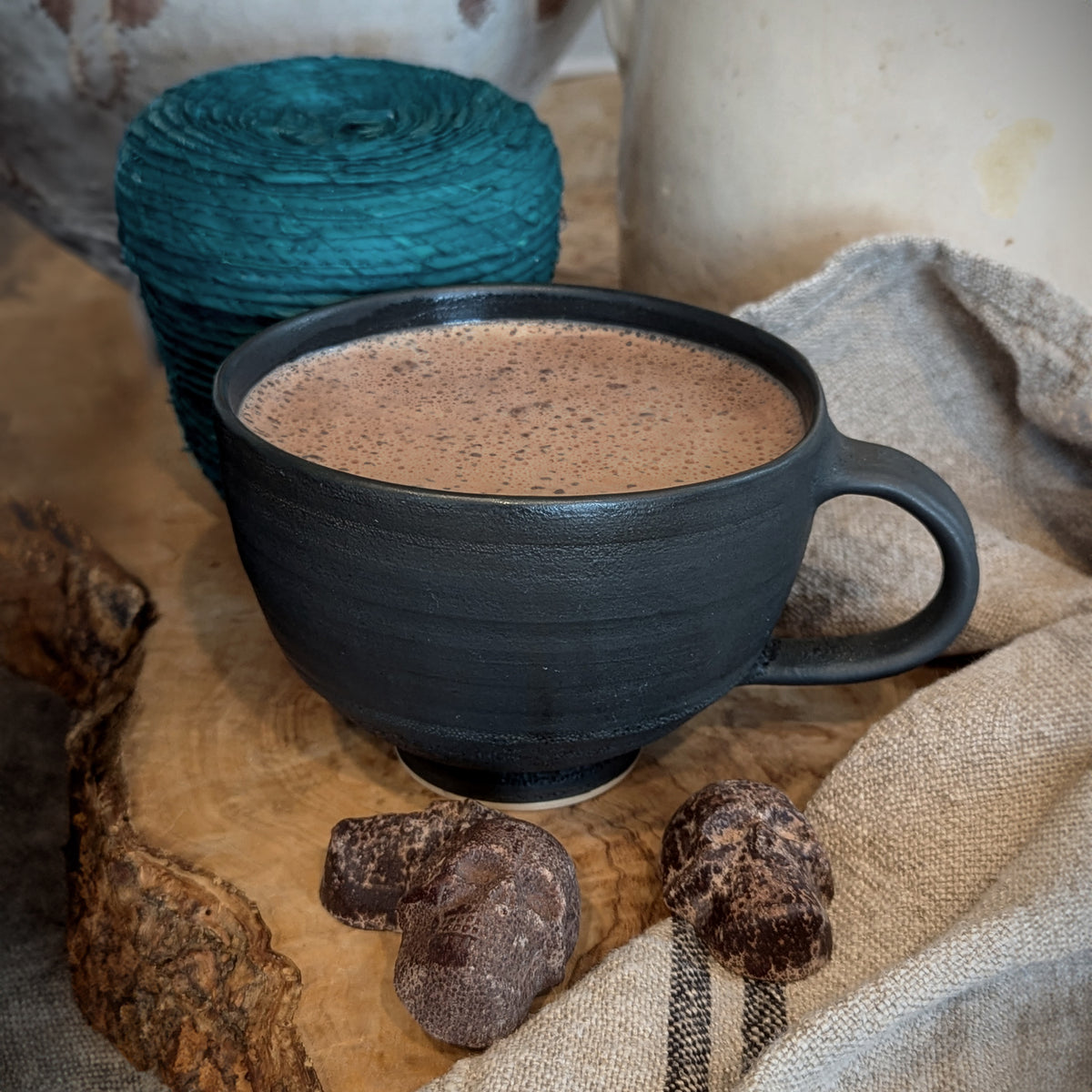 a rustic countertop setting of a finished frothy Traditional Mexican Hot chocolate in a charcoal mug made with the shown cinnamon chocolate skulls . The teal woven basket in the background with a linen kitchen towel