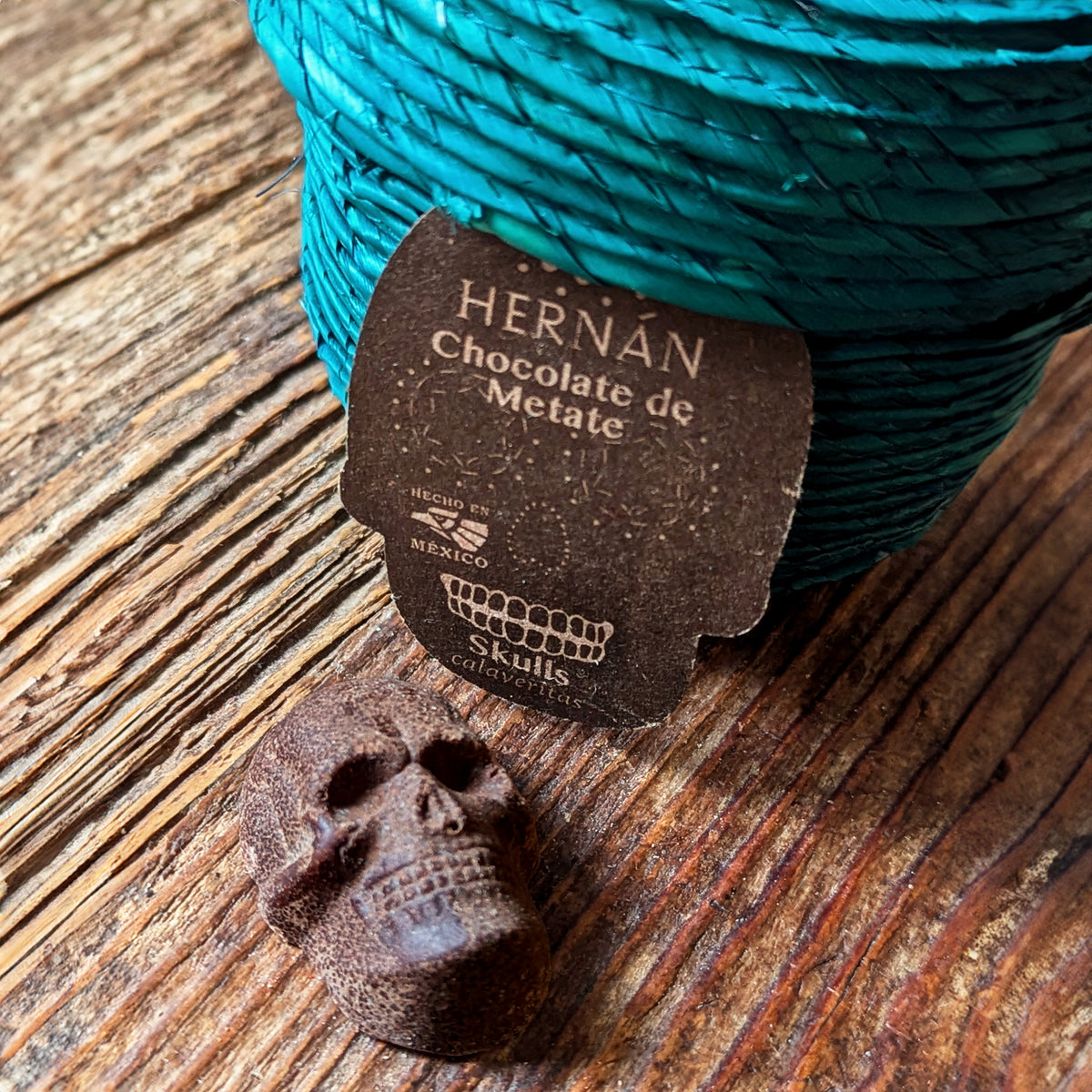Close-up of a single Mexican cinnamon chocolate skull showing molded detail with section of teal palm woven basket with skull shaped label, all resting on a resting on a rustic wooden surface.