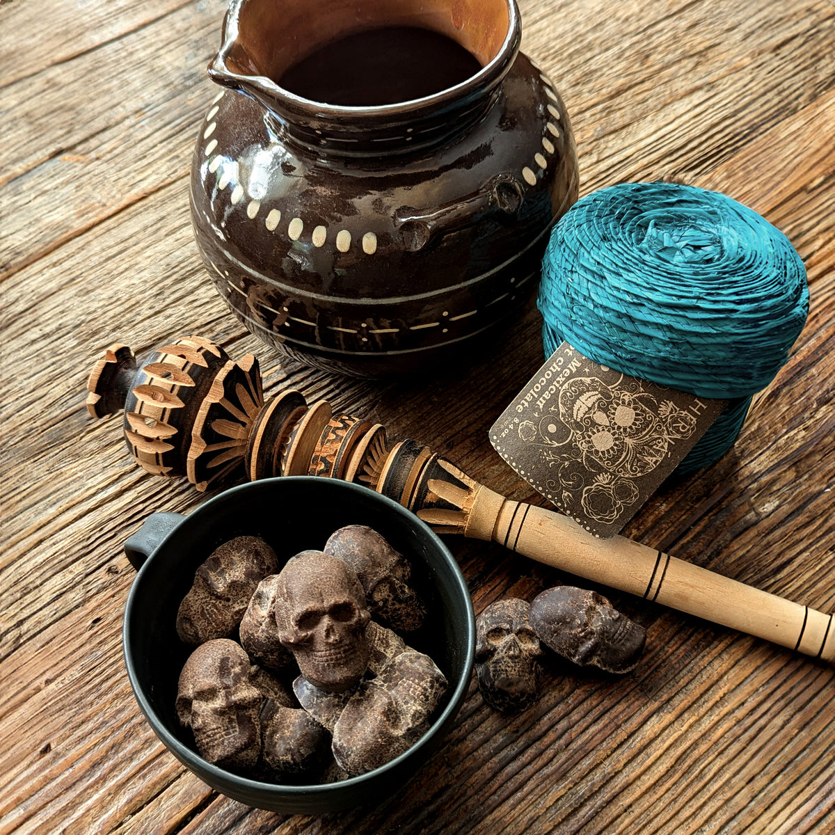 Cinnamon chocolate skulls in a black mug next to a clay olla de barro pot , wooden molinillo frother, and with it's teal palm leaf woven basket styled for traditional Mexican hot chocolate.