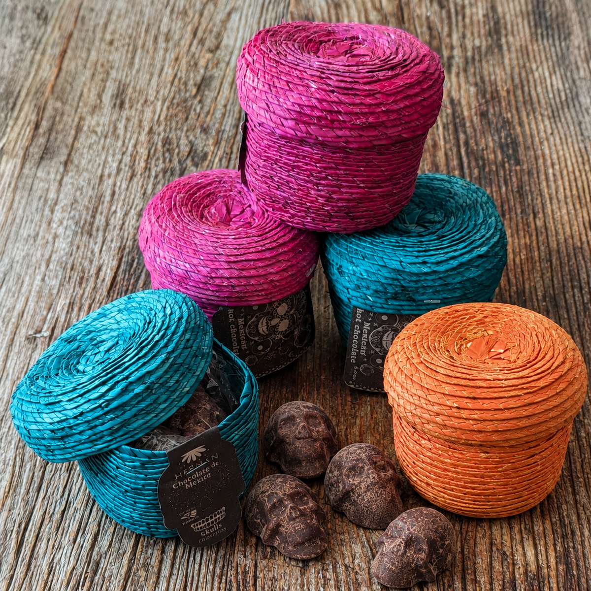 stacked image of 4 palm baskets, two teal and two magenta, each filled with handmade Mexican chocolate skulls, displayed on a rustic wooden surface.