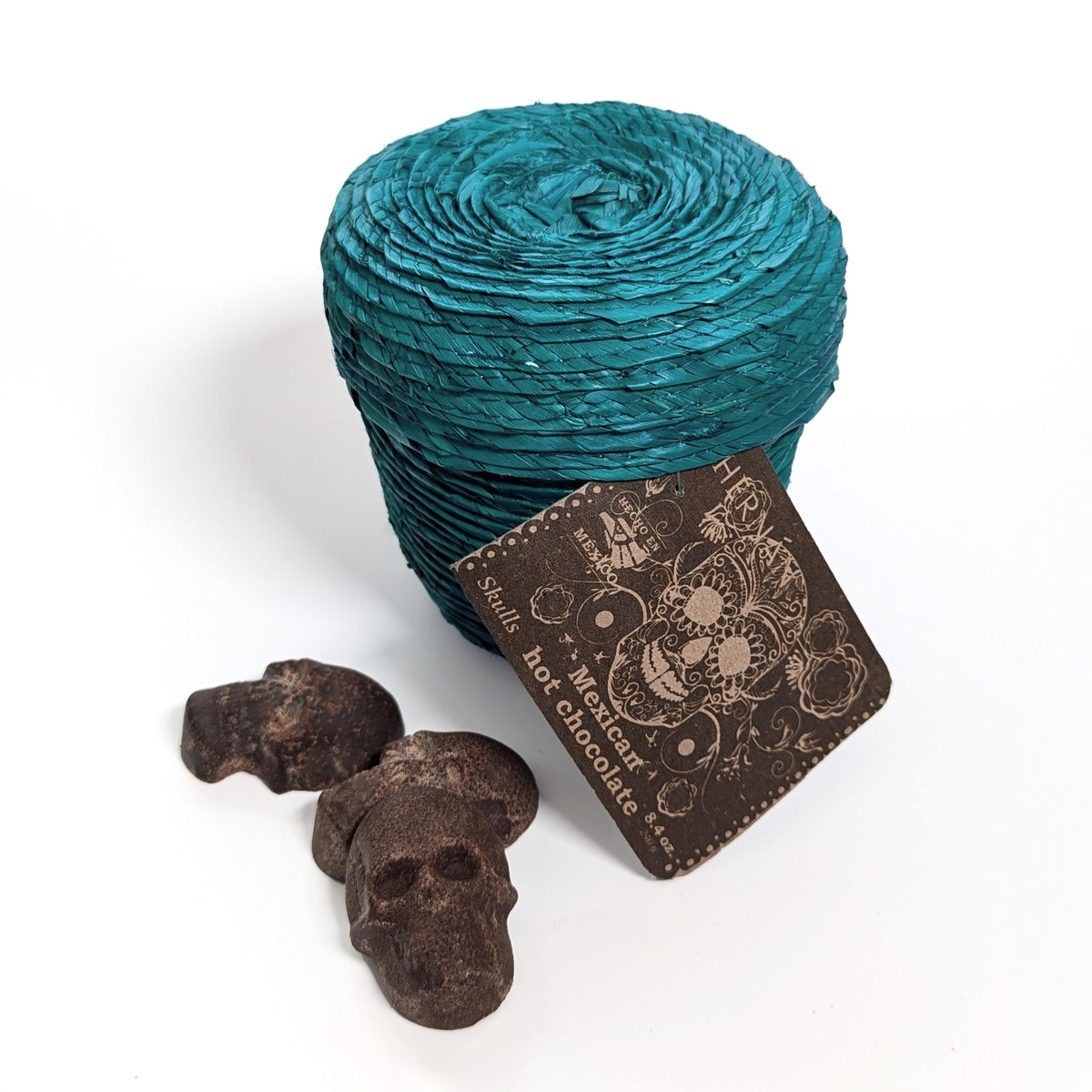 hand-sculpted Mexican cinnamon chocolate skulls arranged next to teal palm leaf woven basket, on a white surface with label