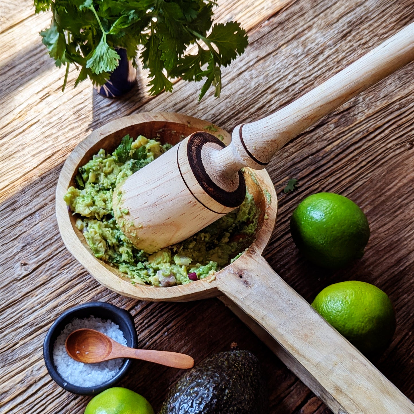 Wooden mortar and pestle with guacamole on a wooden surface with limes and cilantro.