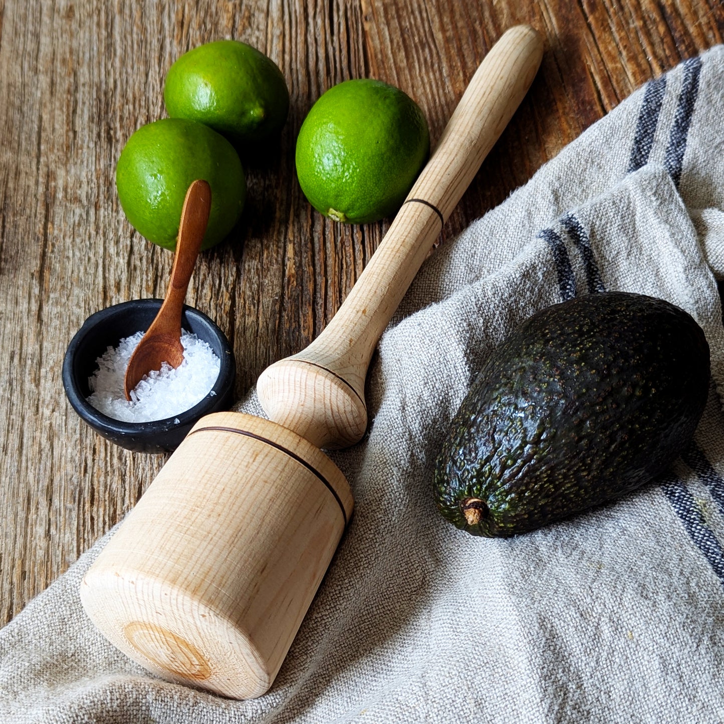 Wooden muddler, avocado, limes and sea salt on a rustic wooden countertop with a linen kitchen towel.