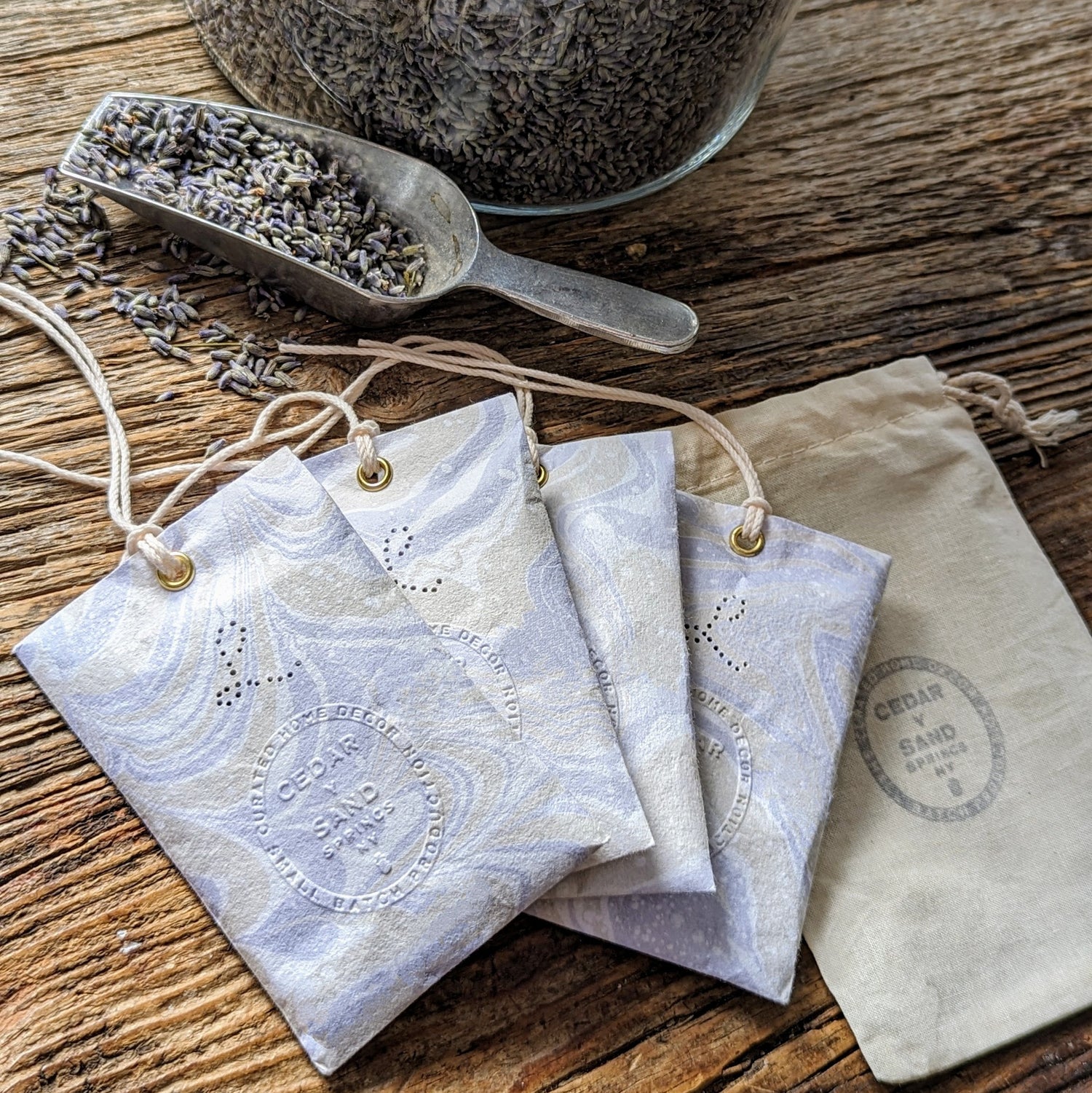 image of the process of filling four lavender hued marble paper lavender sachets with dried lavender buds in a jar and filling scoop .