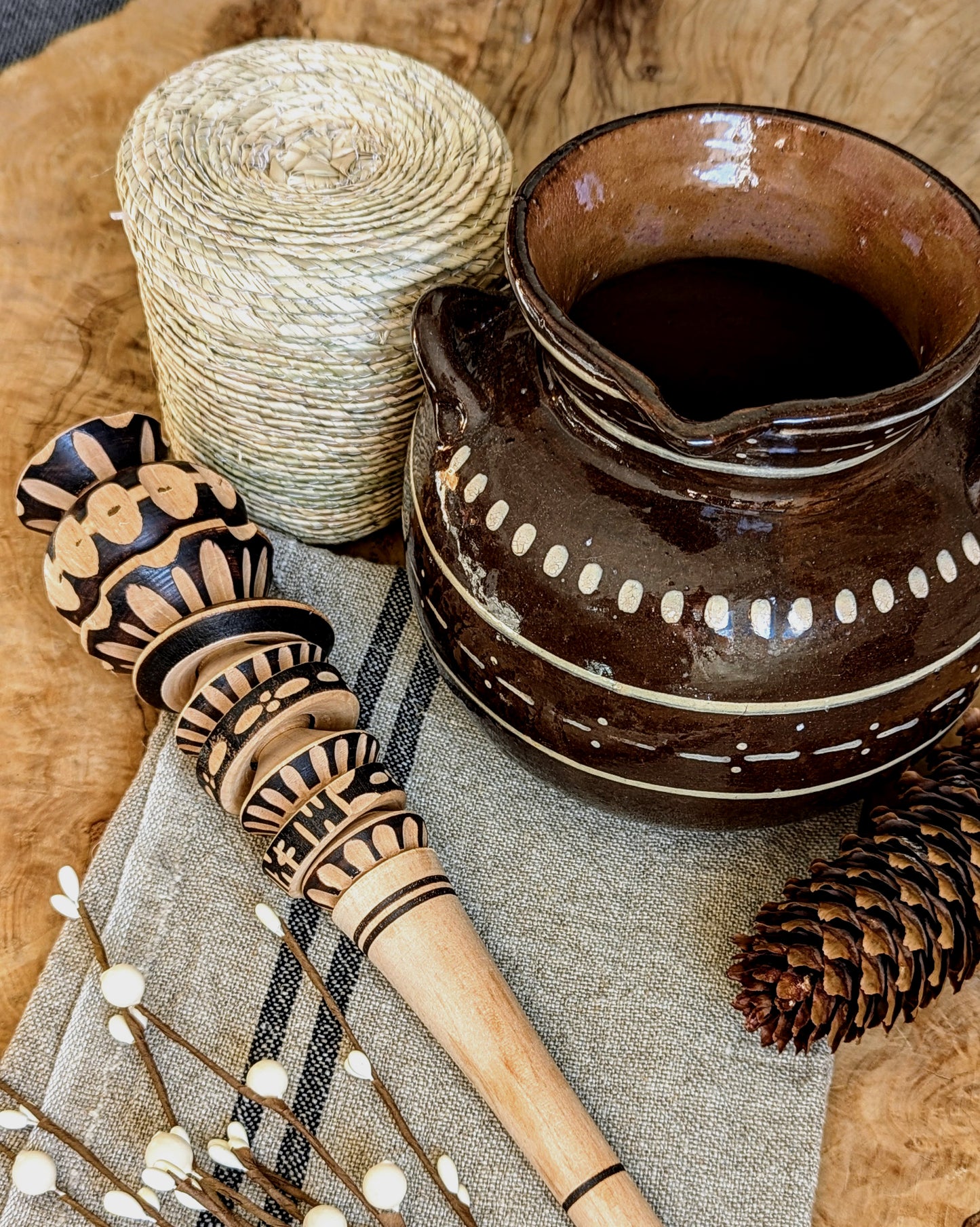 A ceramic Mexican hot chocolate pot with a palm leaf woven basket filled with chocolate bolitas, a wooden frother, displayed on a rustic linen kitchen towel