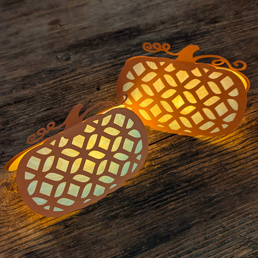 Pair of Pumpkin Lattice Notecards glowing as mini lanterns with Autumn Glow Tealights inside, creating a warm flickering light across a wooden surface.