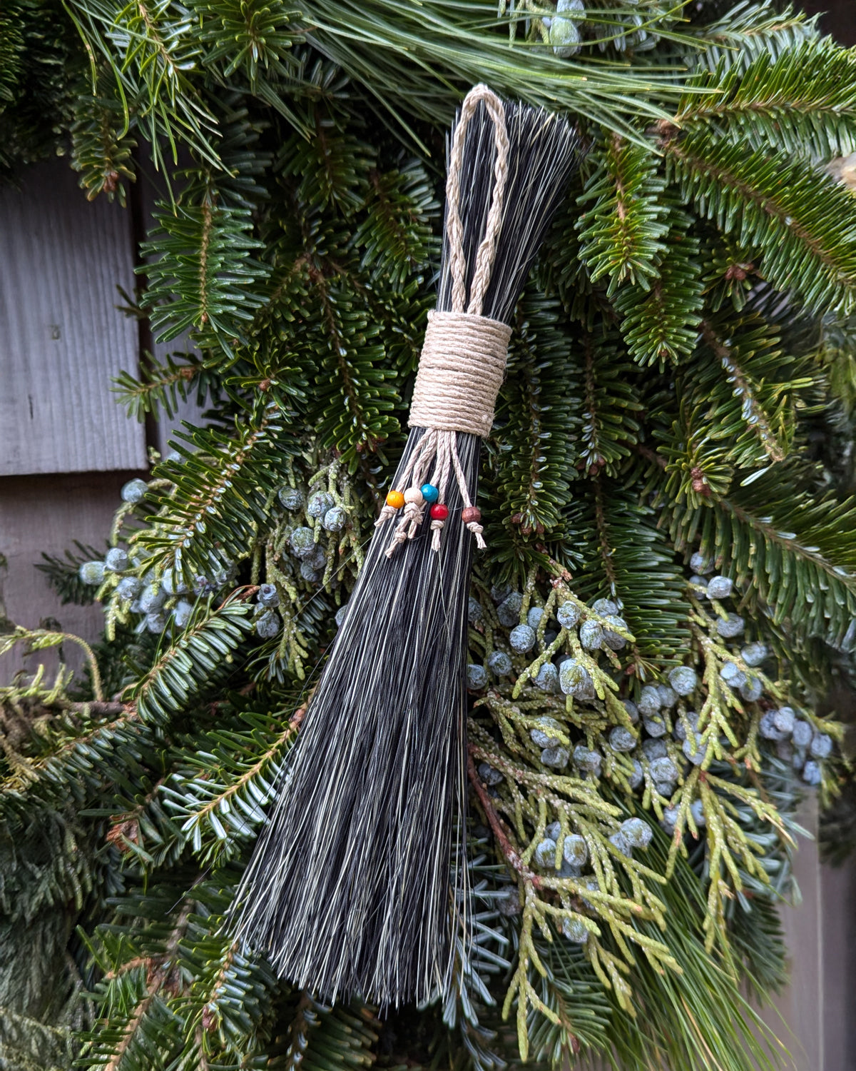 Mixed Agave Slim Table Broom with natural twine handle and colorful bead accents, displayed against a backdrop of fresh evergreen branches and juniper berries, evoking a rustic, seasonal setting.