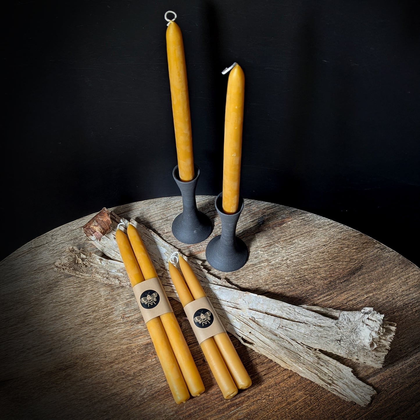 Pure beeswax candlesticks styled in black ceramic holders with additional wrapped candle pairs on a wooden table, offering a warm, rustic home atmosphere.