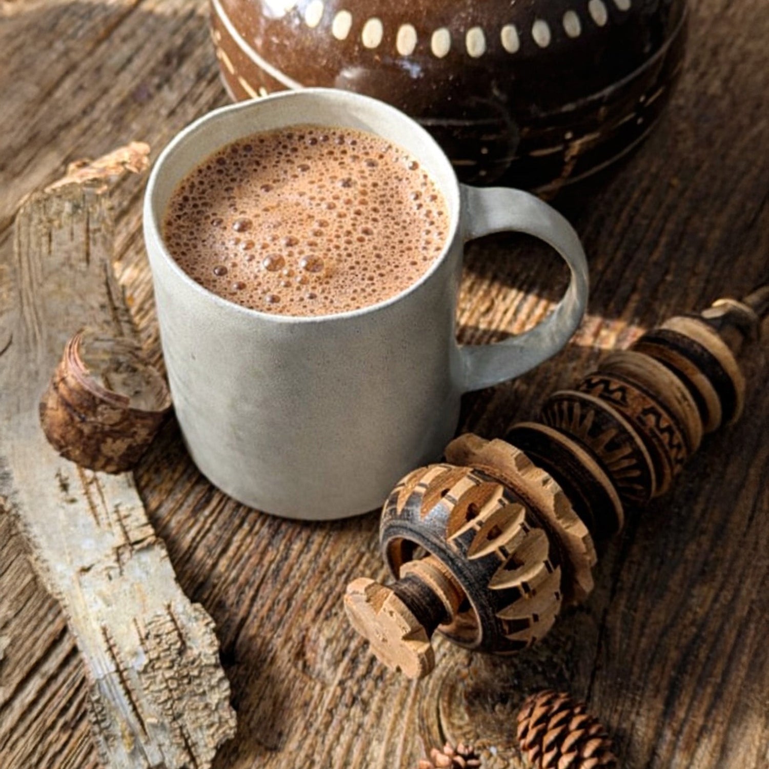 A handcrafted ceramic mug filled with warm cocoa sits on a weathered wooden surface beside a carved wooden molinillo, evoking a quiet winter moment.