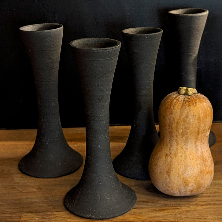 A set of four handcrafted black clay candle taper holders by Jordan B Ceramics displayed on a wooden surface against a dark backdrop. Their tall, sculptural forms stand beside a small tan butternut squash, creating a warm, seasonal composition.