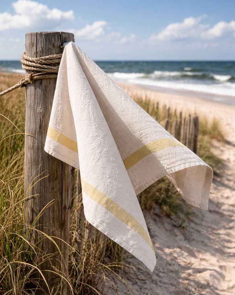 Natural linen towel draped over a weathered wooden post along a coastal dune path overlooking the ocean, featured in The Long Light summer collection by CEDAR Y SAND.