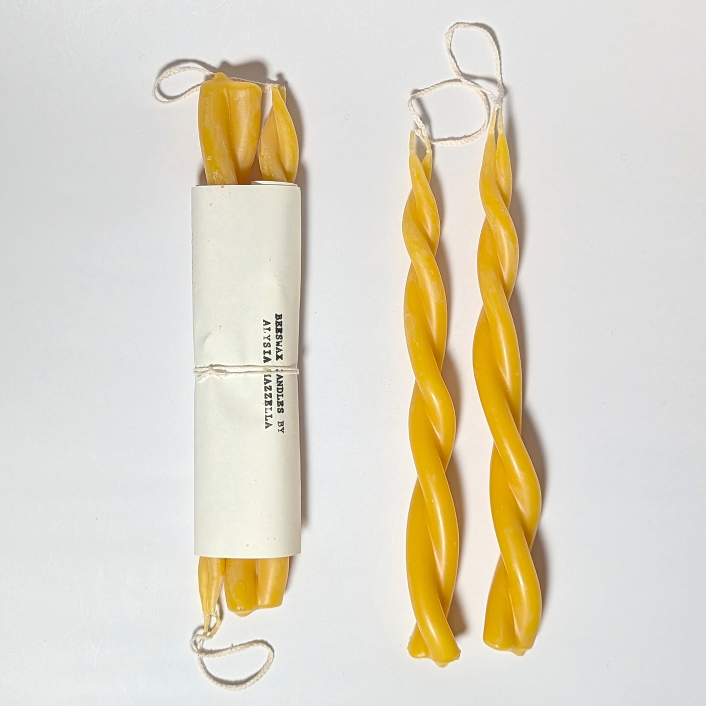 A pair of Twin Flame beeswax candles beside a wrapped bundle labeled “Beeswax Candles by Alysia Mazzella,” emphasizing the handmade detail and pure beeswax texture.