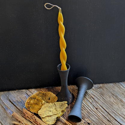 A single Twin Flame beeswax candle displayed in a black ceramic taper holder, surrounded by dried florals, showcasing contrast between matte clay and golden wax.