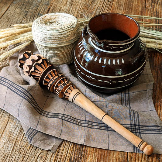 A hand-carved wooden molinillo frother rests on a striped linen kitchen towel beside a rustic olla de barro ceramic pot and palm woven basket filled with chocolate bolitas styled on a natural wood surface.