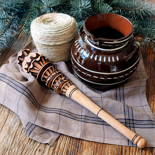 A hand-carved wooden molinillo frother rests on a striped linen kitchen towel beside a rustic olla de barro ceramic pot and palm woven basket filled with chocolate bolitas styled on a natural wood surface with evergreens.