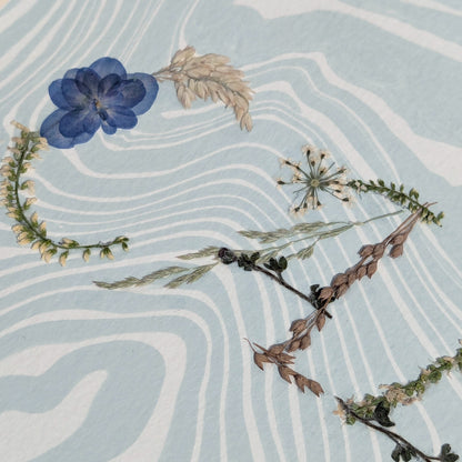 Detailed view of pressed floral word “CALM” highlighting blue botanicals on marbled Suminagashi paper