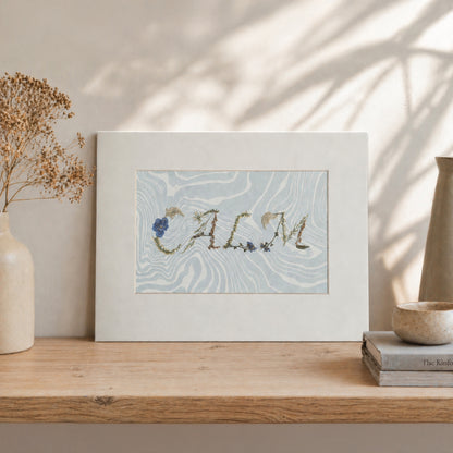 Pressed floral word “CALM” on blue Suminagashi marbled paper, matted botanical artwork styled in a neutral interior setting