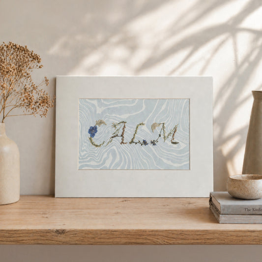 Pressed floral word “CALM” on blue Suminagashi marbled paper, matted botanical artwork styled in a neutral interior setting