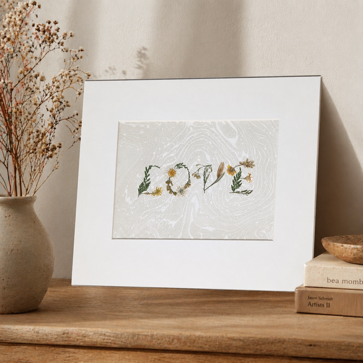 Framed “LOVE” botanical artwork made from pressed flowers on a Suminagashi marbled paper, styled on wooden surface with ceramic vase, dried florals, and stacked books.