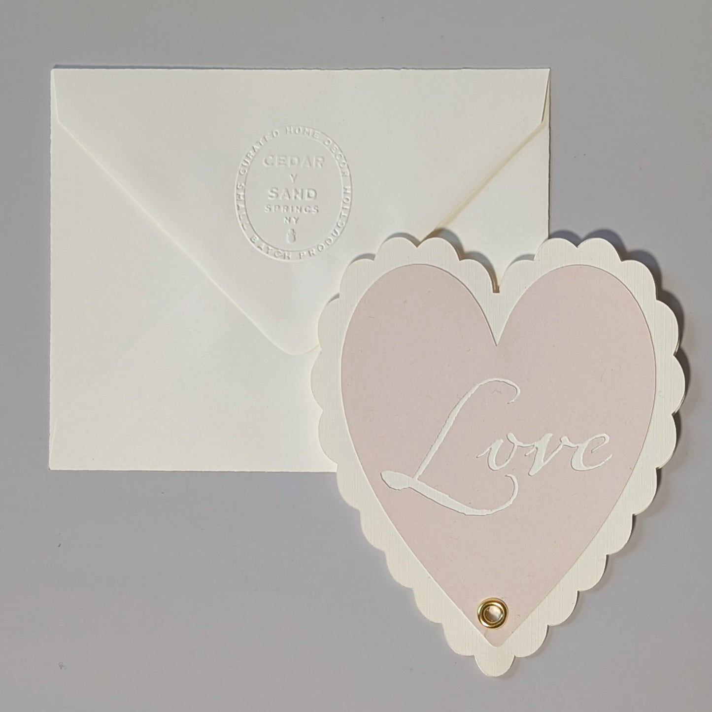 Front view of sculptural heart notecard with cut-out “Love” sentiment, brass rivet detail, and embossed Cedar y Sand envelope.