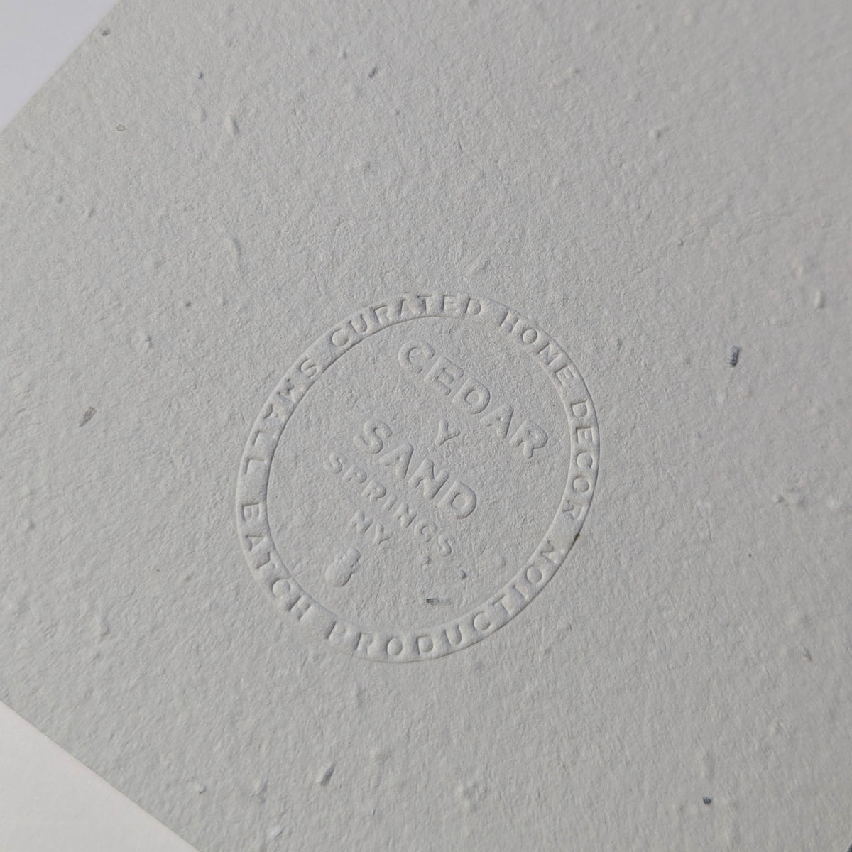 Close-up of the blind-embossed CEDAR Y SAND insignia on textured handmade plantable wildflower seed paper.