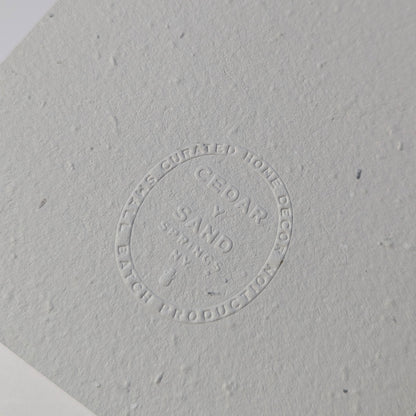 Close-up of the blind-embossed CEDAR Y SAND insignia on textured handmade plantable wildflower seed paper.