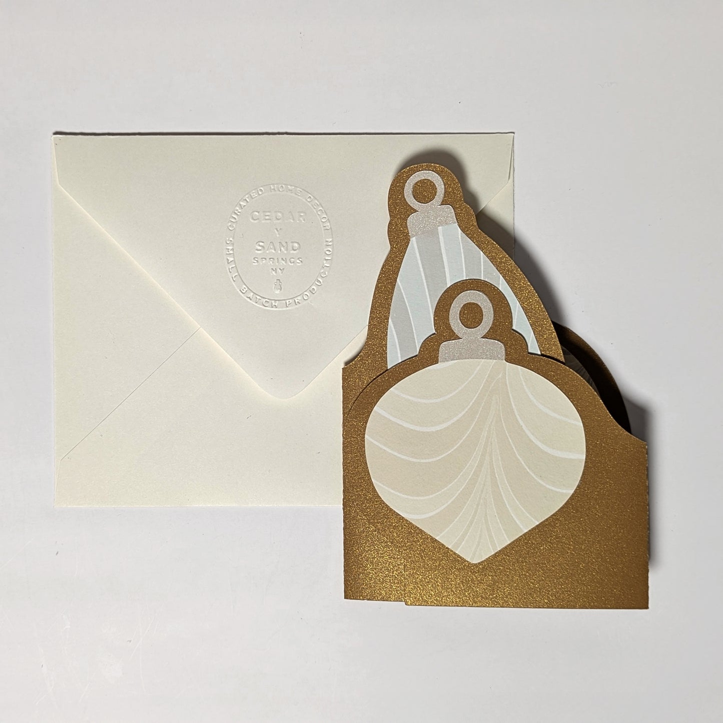 The Suminagashi trifold ornament notecard folded flat, revealing the front ornament shape layered with marbled paper, paired with a cream envelope embossed with the CEDAR Y SAND seal.
