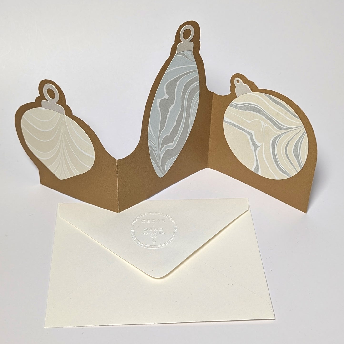 A gold trifold ornament notecard standing open on a white surface, featuring three Suminagashi-marbled ornament panels in soft neutral tones with an embossed CEDAR Y SAND envelope below.