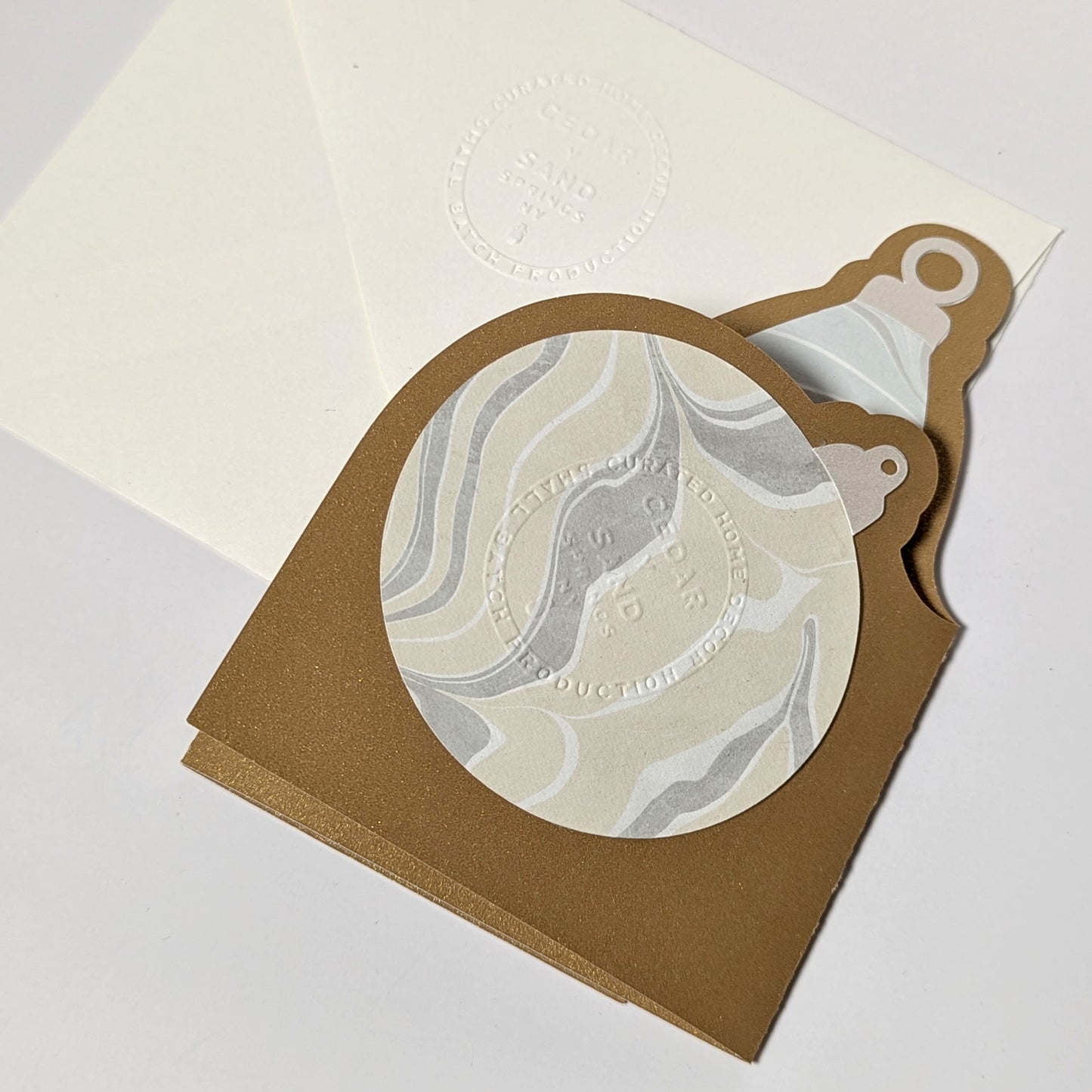 The back of the Suminagashi trifold ornament notecard, showing the marbled circular ornament panel with logo emboss and the CEDAR Y SAND embossed envelope beneath.
