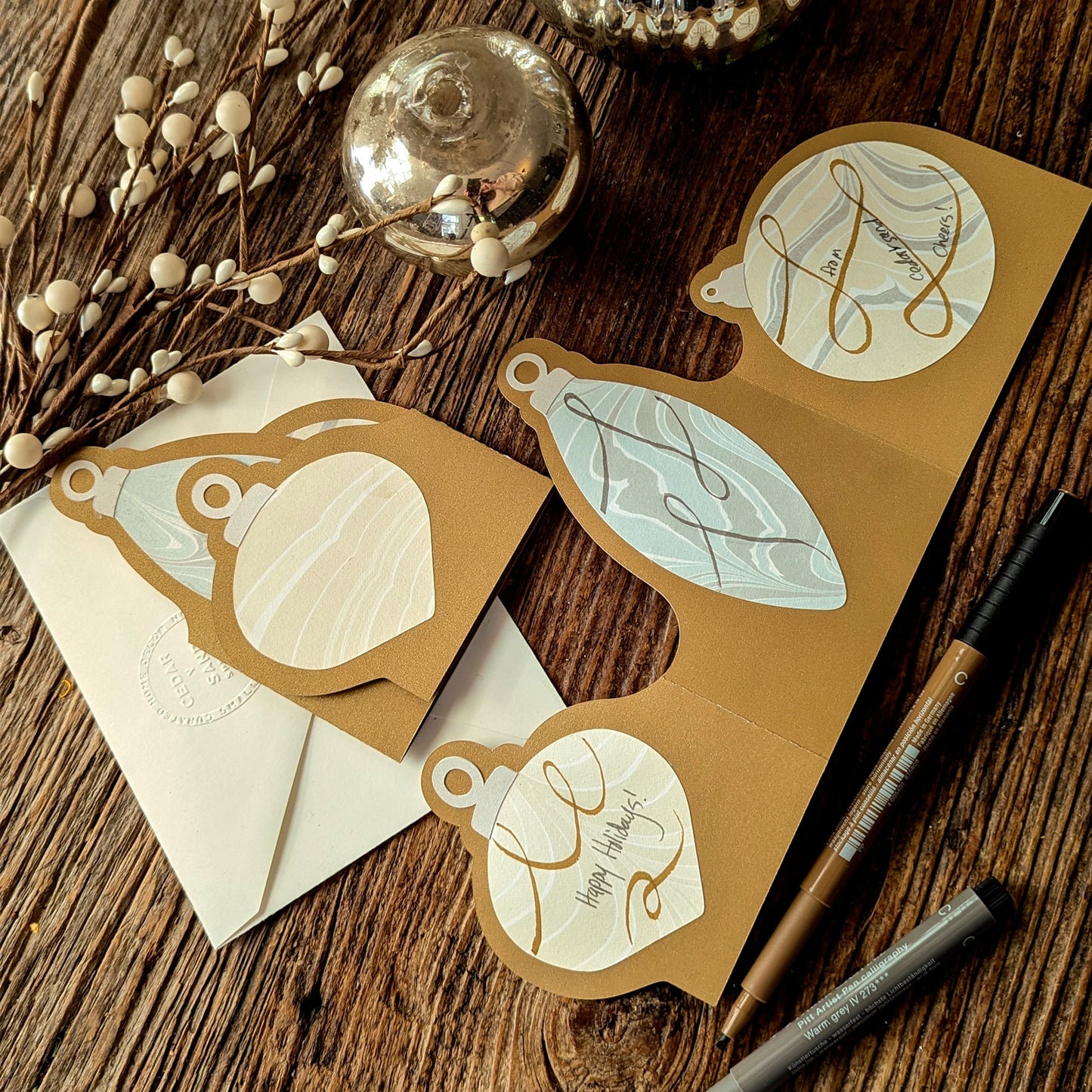 Two Suminagashi trifold ornament notecards—one fully embellished with scroll details, the other blank—displayed with holiday ornaments and white berry stems on a rustic wooden table.