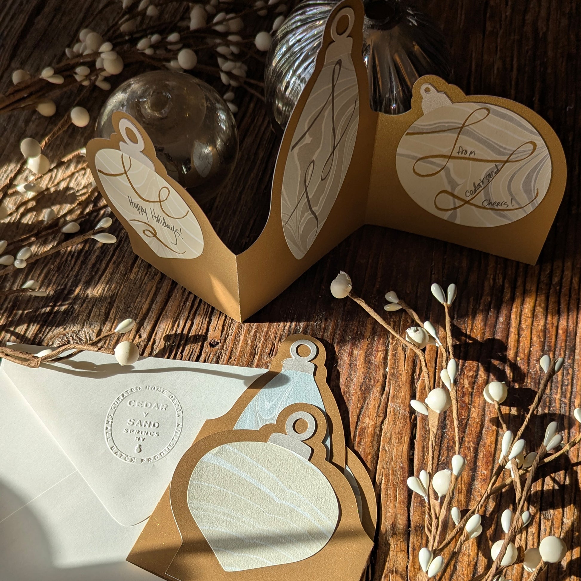 A sunlit trifold ornament notecard displayed on a rustic wooden surface with metallic holiday ornaments and berry branches, showing hand-drawn ribbon-style embellishments on each marbled ornament panel. Card mot sold with added embellishment.