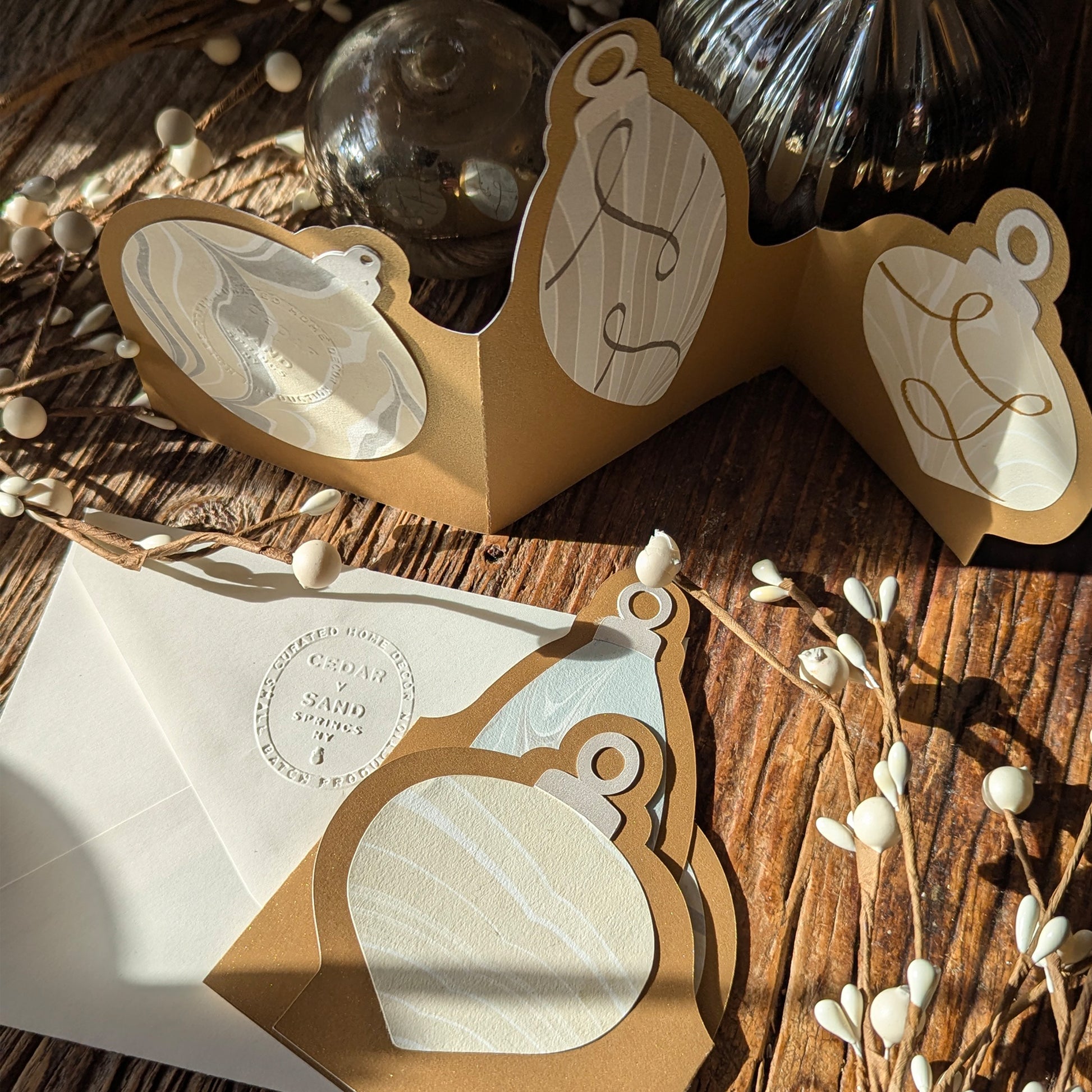 A warm, seasonal scene with the Suminagashi trifold ornament notecard backside partially opened, featuring gold marker scroll embellishments and the handwritten message “Happy Holidays,” surrounded by holiday décor. *Card not sold with added embellishment