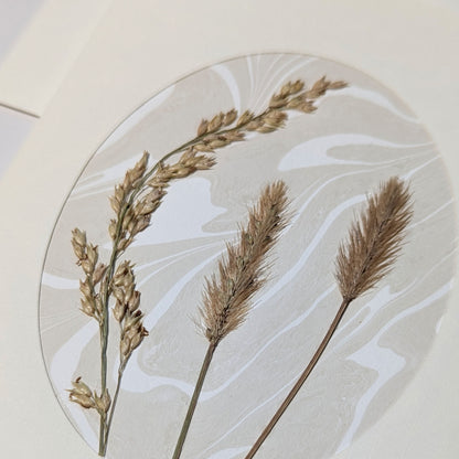 Close-up of Walking Dunes Straw pressed flora notecard with two feathery golden grass plumes and an arching seed stem over hand-marbled Suminagashi paper.