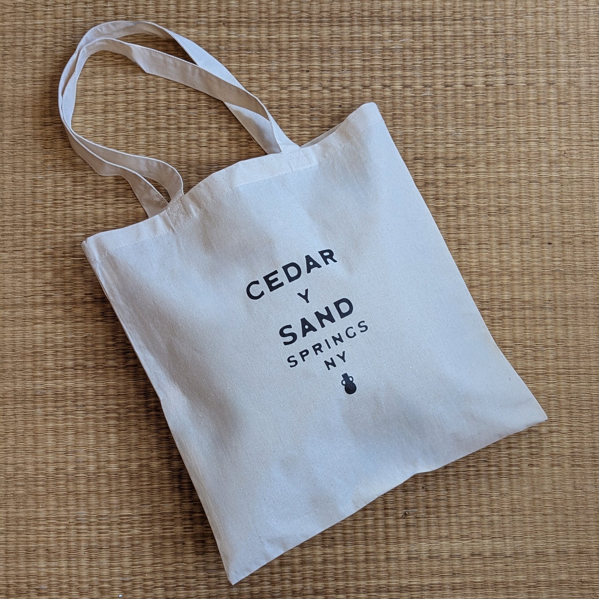 A natural canvas tote bag printed with the CEDAR Y SAND Springs NY logo lies flat on a woven mat background.
