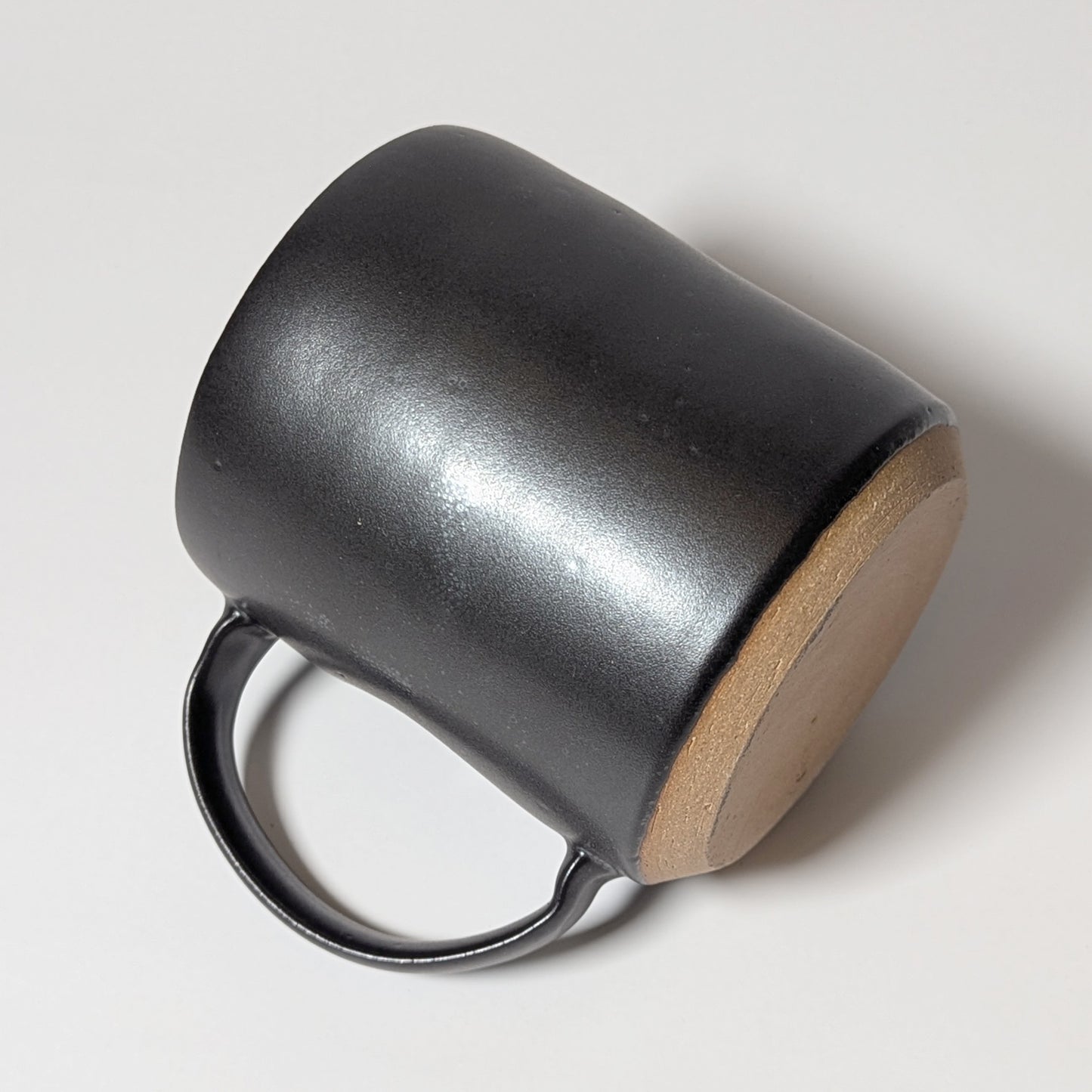 Hand-thrown black stoneware mug resting on its side, showing raw clay base and curved ergonomic handle