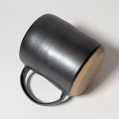 Hand-thrown black stoneware mug resting on its side, showing raw clay base and curved ergonomic handle