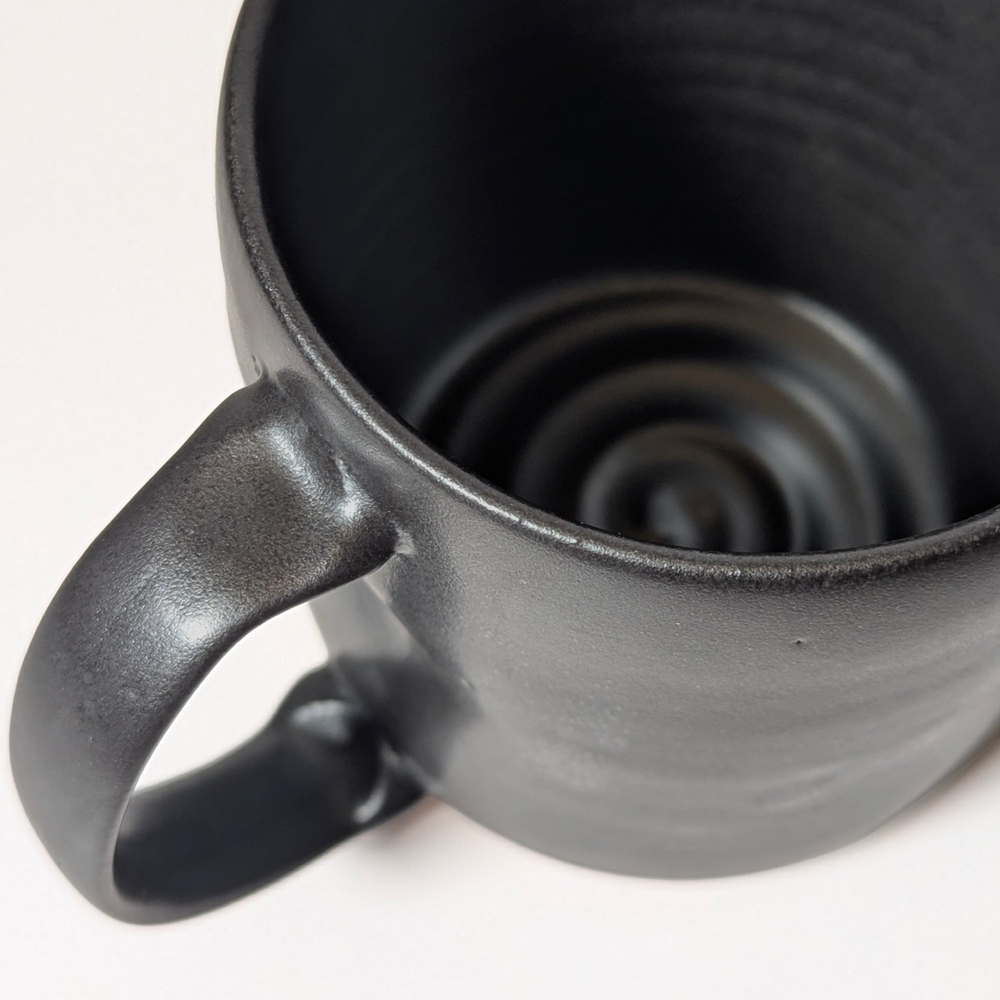 Close-up of smooth extruded handle on black stoneware mug with subtle glaze texture. Shows thumb indent on top of handle for a better grip