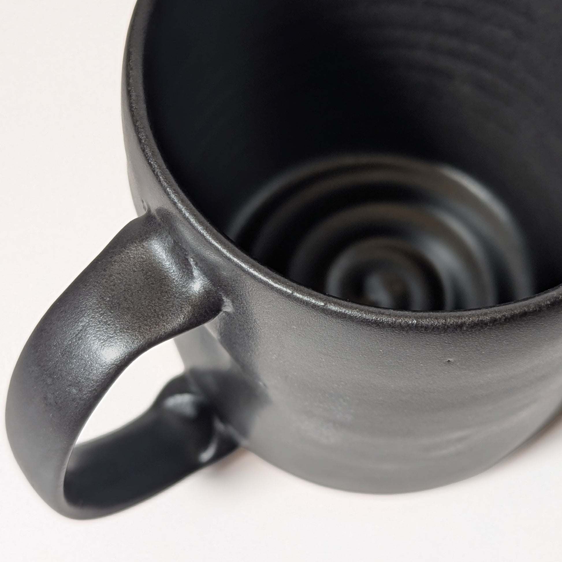 Close-up of smooth extruded handle on black stoneware mug with subtle glaze texture. Shows thumb indent on top of handle for a better grip
