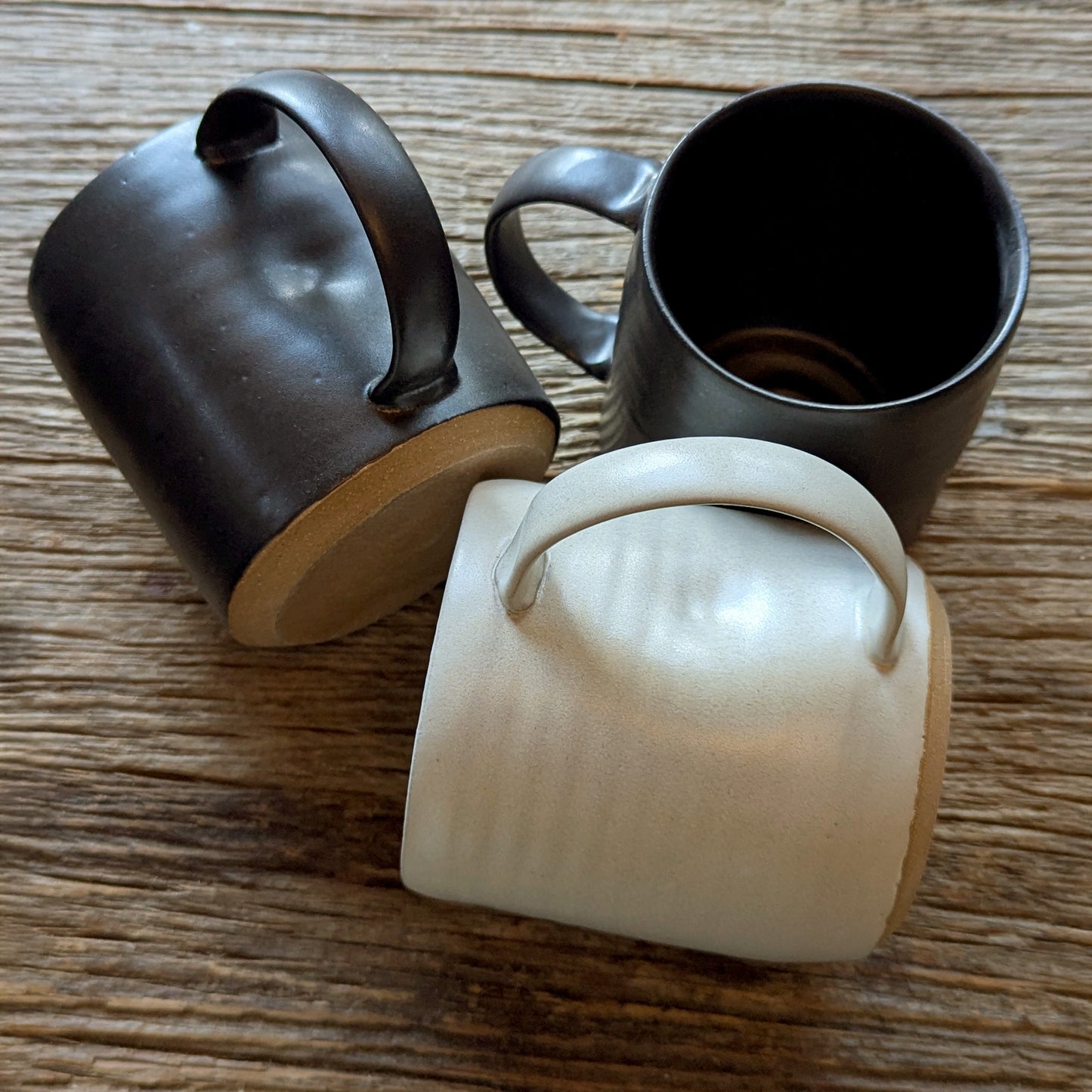 Black and off-white handmade stoneware mugs shown from above and side, highlighting raw clay bases and smooth handles