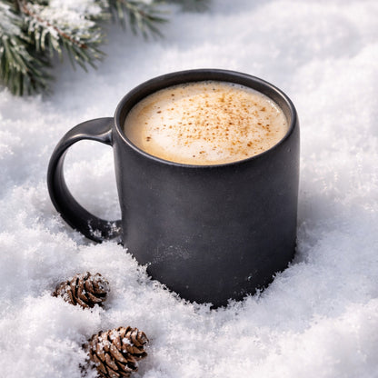 Black stoneware mug filled with frothy coffee resting in fresh snow, with pinecones and evergreen branches nearby