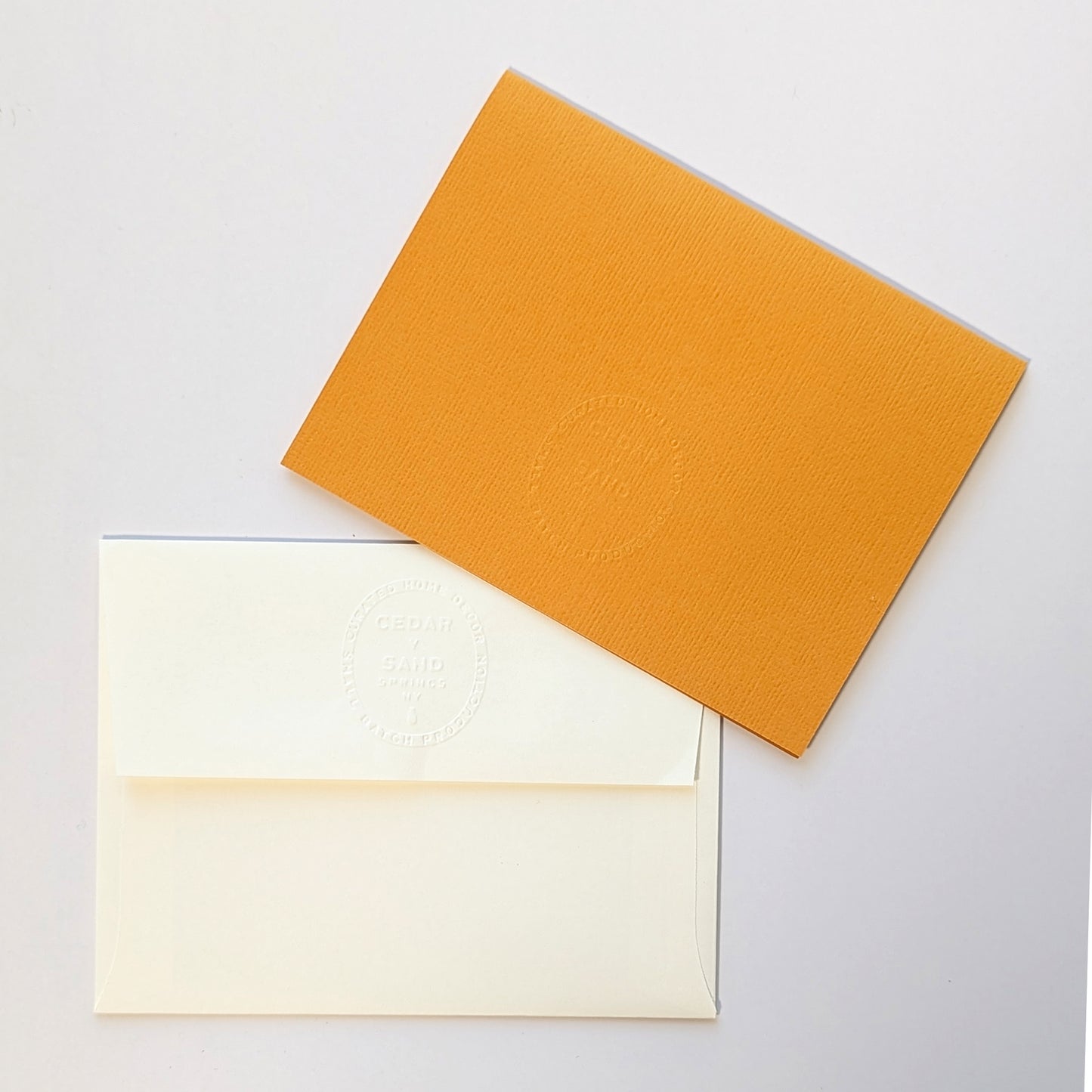 Reverse side of butternut colored notecard and natural colored envelope, both with the CEDAR Y SAND embossed insignia.