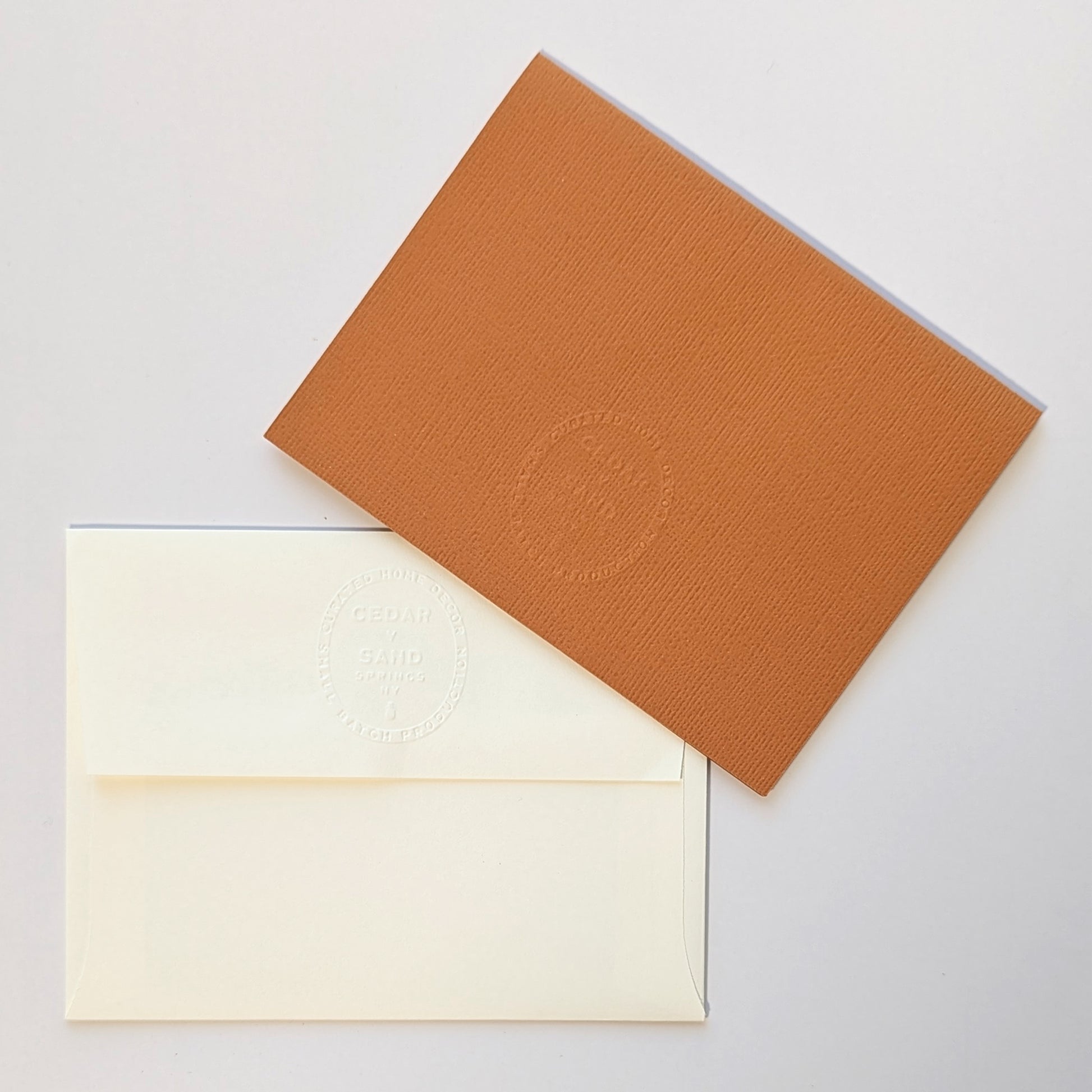 Reverse side of pumpkin colored notecard and natural colored envelope, both with the CEDAR Y SAND embossed insignia.