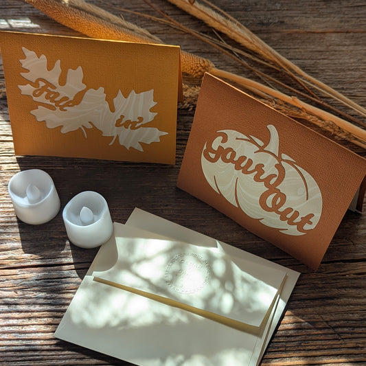Warm, moody scene of “Gourd Out & Fallin’” lantern cards lit with tealights, styled on a wooden surface for a cozy autumn atmosphere.